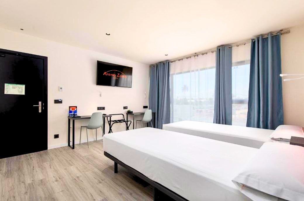 habitación de hotel en valencia donde se ve el interior de la habitación: dos camas, escritorio, ventana con cortinas y la puerta de acceso